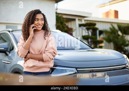 Lächelnde Frau, die über das Mobiltelefon spricht, während sie sich auf das Auto im Hof lehnt Stockfoto