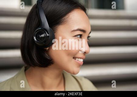 Nahaufnahme Porträt von lächelnden asiatischen Mädchen in Kopfhörern, hört Musik im Freien, schauen glücklich. Stockfoto