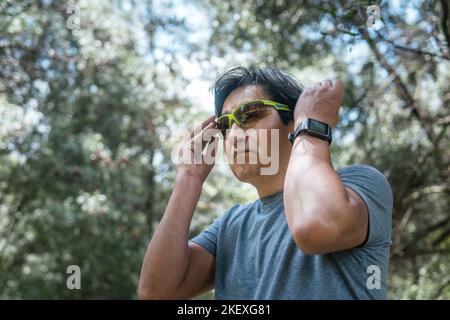 Ein reifer Latino-Mann mit einer Smartwatch, der mitten im Wald eine Laufbrille aufsetzt. Sport- und Technologiekonzept Stockfoto