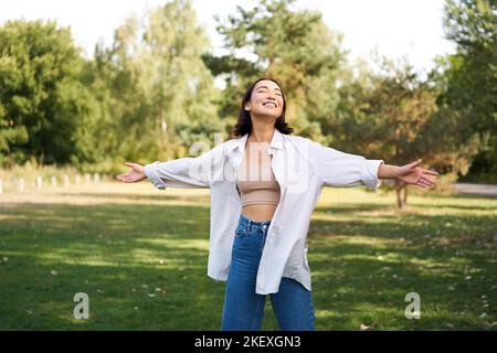 Sorgloses asiatisches Mädchen, das im Park lacht und tanzt, genießt den sonnigen Sommertag, hebt die Hände hoch und atmet frische Luft Stockfoto