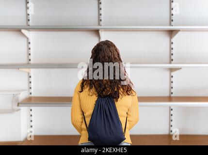 Rückansicht einer jungen Frau, die in einem Supermarkt vor einem leeren Regal steht Stockfoto