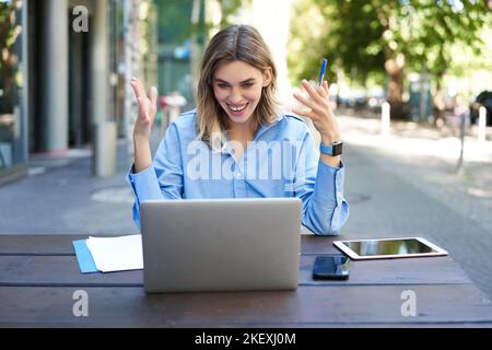 Junge Geschäftsfrau, nehmen an einer Online-Videokonferenz Teil, schreiben, arbeiten, Notizen in ihren Dokumenten machen, draußen an der frischen Luft sitzen Stockfoto