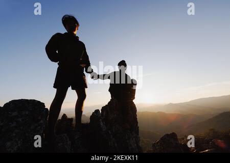 Silhouette eines Wanderers, der sich beim Sonnenuntergang gegenseitig beim Aufstieg auf einen Berg unterstützt. Menschen helfen und, Team arbeiten Konzept. Stockfoto