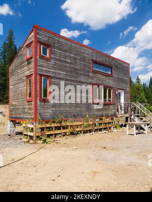 Rustikales Cottage-Stil 11,5 x 32 Fuß Minihaus Fassade mit geneigtem Dach und horizontaler Hemlock Holzverkleidung und rot verzierten Fenstern im Sommer. Stockfoto