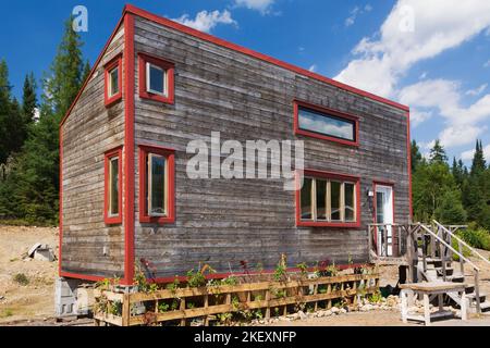 Rustikales Cottage-Stil 11,5 x 32 Fuß Minihaus Fassade mit geneigtem Dach und horizontaler Hemlock Holzverkleidung und rot verzierten Fenstern im Sommer. Stockfoto