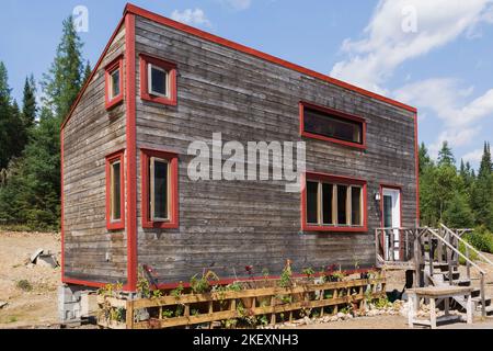 Rustikales Cottage-Stil 11,5 x 32 Fuß Minihaus Fassade mit geneigtem Dach und horizontaler Hemlock Holzverkleidung und rot verzierten Fenstern im Sommer. Stockfoto