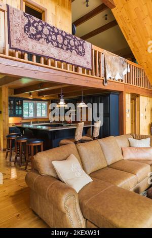 Braunes Ledersofa im Wohnzimmer mit Blick auf Küche und Mezzanin in einem gefrästen Cottage-Stil flaches Blockhaus. Stockfoto