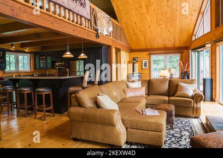 Braunes Ledersofa im Wohnzimmer mit Blick auf Küche und Mezzanin in einem gefrästen Cottage-Stil flaches Blockhaus. Stockfoto