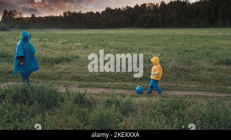 Frau spielt Ball mit kleinem Kind. Sonnenuntergang, bewölktes Wetter, Menschen in Regenmänteln Stockfoto