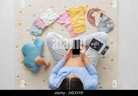 Schwanger Frau, die Telefon-App zum Einkaufen, telemedizinische Lesen über Schwangerschaft oder andere. Schwarzer Bildschirm mit Kopierraum, Babykleidung und Spielzeug Stockfoto