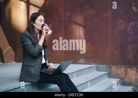 Junge attraktive Geschäftsfrau, die Kaffee trinkt und mit einem Laptop arbeitet, während sie draußen auf einer Treppe sitzt. Online arbeiten, online arbeiten. Weiblich Stockfoto