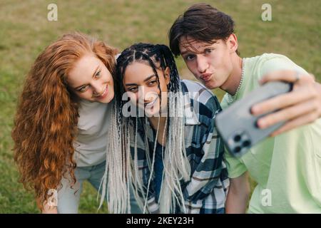 Drei junge, glücklich lächelnde kaukasische Teenager, die im Sommer Selfie im Park machen und verrückt werden und lachen. Sonnenuntergang Licht.Glückliche und positive Emotionen. Stockfoto
