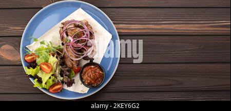 Gegrillter Kebab mit Soße und Gemüse auf einem Teller auf einem Holzhintergrund, Draufsicht Stockfoto