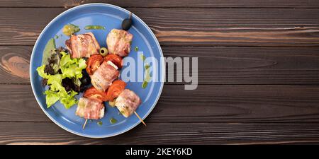 Gegrillte Spieße mit Lachs, eingewickelt in einen Speck mit Tomaten und Salat auf Holzboden, Draufsicht. Banner. Stockfoto