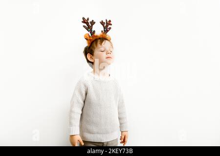 Portrait des kleinen Jungen im Pullover mit weihnachtlichen Stirnband auf hellem Wandhintergrund Stockfoto