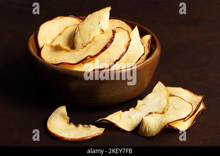 Getrocknete Apfelscheiben in einer braunen Holzschüssel neben verschütteten getrockneten Apfelscheiben auf dunklem Holzhintergrund. Stockfoto