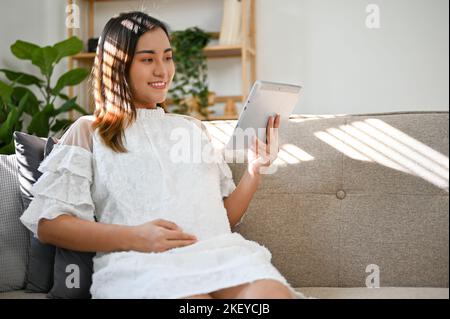 Glückliche und schöne asiatische Schwangere, die in ihrem Wohnzimmer ruht, ihren Bauch berührt und Babypflege auf ihrem Tablet beobachtet. Mutterschaft Konzept Stockfoto