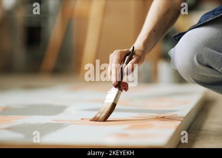 Selektiver Fokus auf die Hand der jungen kreativen Frau mit Pinsel schaffen abstrakte Malerei auf Leinwand, während hocken auf dem Boden Stockfoto