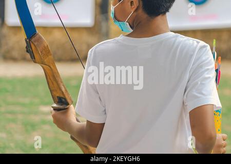 Rückansicht der asiatischen Jungen tragen Gesichtsmaske zielt Bogenschießen Bogen und Pfeil zu bunten Ziel im Schießstand während des Trainings und des Wettbewerbs. Übung und Co Stockfoto