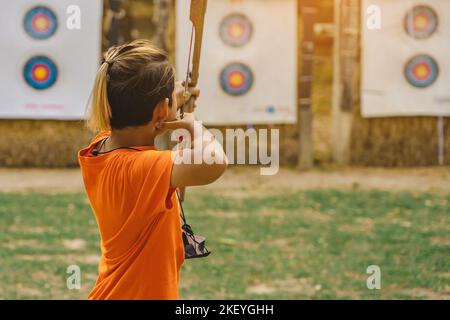 Rückansicht der asiatischen Jungen tragen Gesichtsmaske zielt Bogenschießen Bogen und Pfeil zu bunten Ziel im Schießstand während des Trainings und des Wettbewerbs. Übung und Co Stockfoto