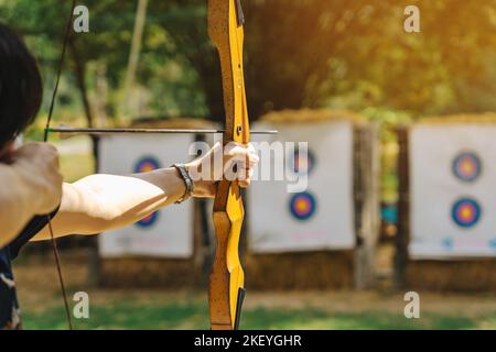 Hände der Frau zielt Bogenschießen Bogen und Pfeil auf bunte Ziel im Schießstand während des Trainings. Bewegung und Konzentration beim Bogenschießen im Freien. Auswahl Stockfoto