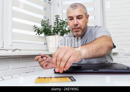 Nahaufnahme eines Mannes, der einen Laptop mit einem Schraubendreher und verschiedenen Werkzeugen auf dem Tisch auseinandernimmt. Der Meister legt kleine Schrauben auf eine Magnetmatte. Stockfoto