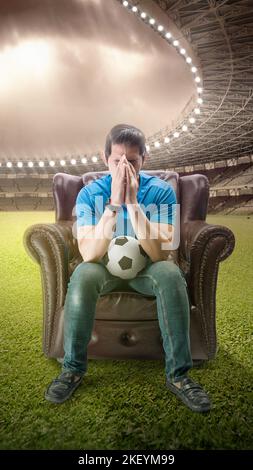 Asiatischer Mann mit dem Ball auf der Couch mit einem enttäuschten Ausdruck im Hintergrund des Fußballstadions Stockfoto