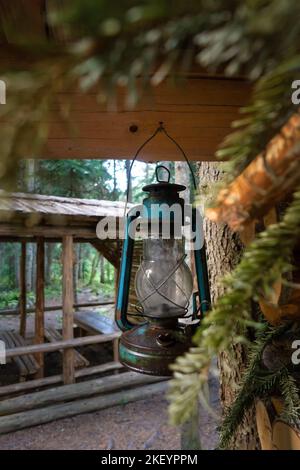 Alte Kerosin-Laterne, die an der Ecke eines Holzblockhauses hängt und neben einem Pavillon im Wald steht. Vertikales Foto Stockfoto