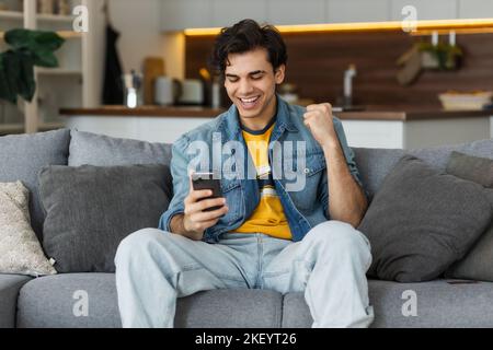 Aufgeregter junger Kaukasusmann, der Erfolg feiert. Junger Mann, der gute Nachrichten mit dem Mobiltelefon liest, das auf dem Sofa sitzt Stockfoto
