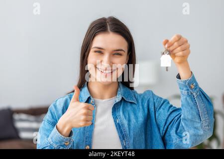 Glückliche junge kaukasische Besitzerin eines neuen Hauses zeigt den Schlüssel zu einem neuen Zuhause, schaut auf die Kamera und lächelt Stockfoto