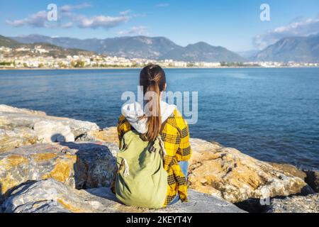 Junge Reisende Frau mit Rucksack sitzt mit dem Rücken zur Kamera auf dem Stein am Meer und schaut in die Ferne Stockfoto