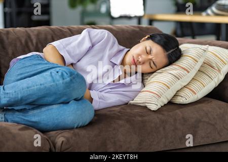 Frau mit Bauchschmerzen, die zu Hause auf der Couch liegt Frau mit starken Krämpfen, die ihren Magen halten Stockfoto
