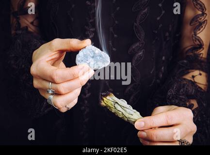 Nahaufnahme einer Frau in schwarzem Spitzenkleid, die den Edelstein eines blauen Himmelsteins aus Kristall reinigt, indem sie das weiße Salbei-Bündel verwischt. Entfernen Sie negative Energie. Stockfoto