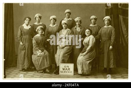 Die Originalpostkarte aus dem Jahr WW1 mit weiblichen Munitionsarbeiterinnen, das Mädchen von Far RH, hat das Abzeichen „im Kriegsdienst“ und vor ihnen ist ein Schild „im Kriegsdienst“ zu sehen. Aus dem Fotostudio von Whyte and Sons, 20 Union St. Glasgow, Schottland, Großbritannien um 1916-1918 Stockfoto