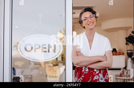 Glückliche Inhaberin des Cafés lächelt der Kamera zu, während sie neben einem offenen Schild am Eingang ihres Cafés steht. Reifer Inhaber von Kleinunternehmen, der c Stockfoto