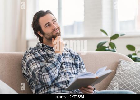 Nachdenklicher, intelligenter Tausendjähriger europäischer Mann im Hemd mit Stoppeln mit Buch oder Tagebuch liest oder träumt auf dem Sofa Stockfoto