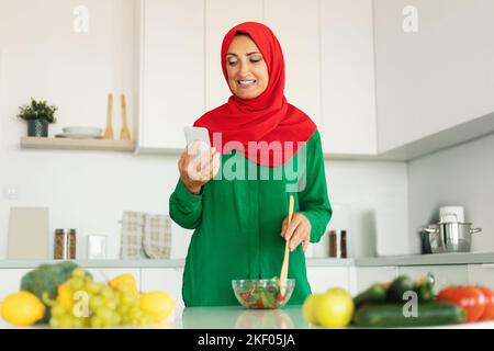 Glückliche reife islamische Frau, die Essen mit Bio-Gemüse zubereitet und im Kücheninnenraum SMS schreibt Stockfoto