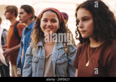 Fröhliches Teenager-Mädchen, das mit einer Gruppe von Demonstranten in der Stadt vor der Kamera lächelt. Multikulturelle Generation z-Aktivisten marschieren für e Stockfoto