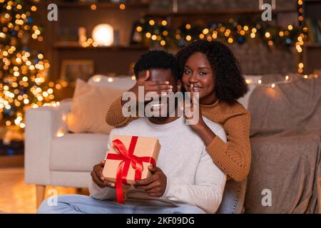 Lächelnde junge afroamerikanische Frau schließt die Augen vor dem Ehemann, zeigt ein Schweigeminde und gibt einen Geschenkkarton Stockfoto