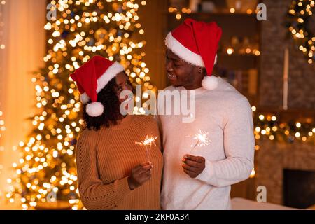 Glückliches junges afroamerikanisches Paar in den Weihnachtsmützen umarmt, hält Wunderkerzen im dunklen Raum Stockfoto