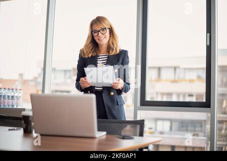 Aufnahme einer selbstbewussten, reifen Geschäftsfrau, die in einem modernen Büro einen Videoanruf abhält und gleichzeitig ein Dokument in der Hand hält. Professionelle weibliche Wea Stockfoto