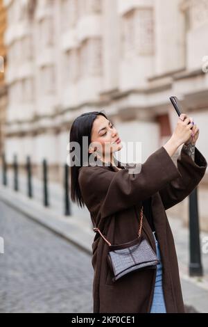 Hübsche junge Frau in braunem Mantel, die Selfie in prag auf unscharfem Hintergrund, Stockbild Stockfoto