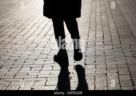 Schwarze Silhouette und Schatten einer einsamen Frau, die auf einer Straße geht. Weibliche Beine in Stiefeln auf gepflastertem Bürgersteig der Stadt Stockfoto