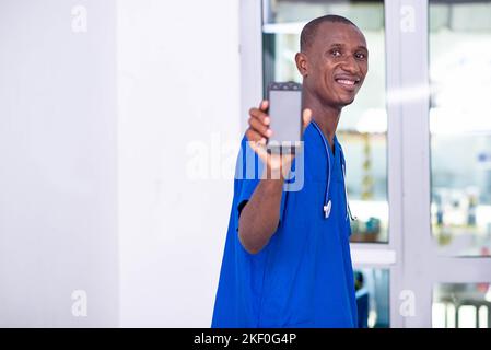 Hübscher junger Arzt in blauer Uniform mit Stethoskop, das den Bildschirm des Mobiltelefons zeigt, während er lächelt. Stockfoto