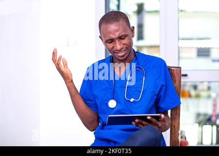 Junge ernsthafte männliche Arzt in blauer Uniform sitzt auf einem Stuhl arbeiten auf digitalen Tablet. Stockfoto