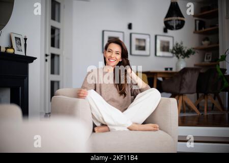 Porträt einer schönen langhaarigen Frau in legerer Kleidung, während sie sich zu Hause in einem Sessel entspannt. Stockfoto