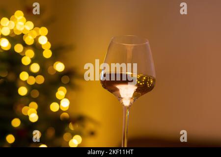 Ein Glas Wein, verschwommene weihnachtslichter auf dem weihnachtsbaum im Hintergrund. Kiefernbaum mit leuchtender Girlande geschmückt, Tasse mit einem Getränk. Stockfoto
