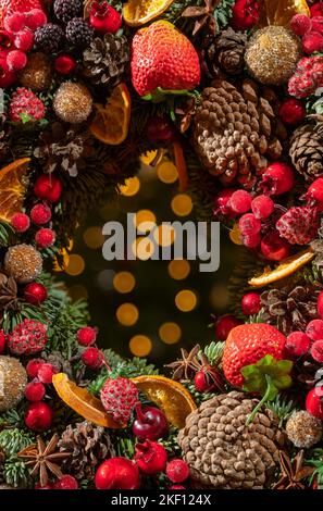 Farbenfroher Kranz im Hintergrund des verschwommenen weihnachtslichts. Girlande verziert mit Früchten und Beeren im Font der verschwommenen Glühbirnen. Stockfoto