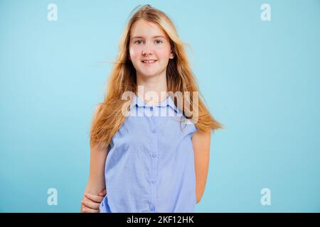 Lose behaarte Teenager-Mädchen stehen mit Händen hinter dem Rücken auf leeren blauen Hintergrund halten. Porträt eines lächelnden Kindes im Schulalter in stilvollem ärmellosen Look Stockfoto