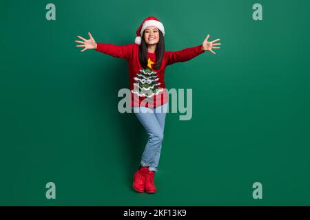 In voller Länge Foto von glänzend aufgeregt Mädchen gekleidet hässlich drucken rot x-Mas Pullover Kopfbedeckung offene Arme wollen Umarmung Sie isoliert grüne Farbe Hintergrund Stockfoto